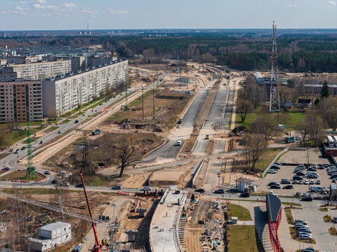 Aerial view of Riga, Latvia, shows Southern Bridge phase 4 work, graded lanes, cranes, heavy machinery, detours, Soviet era blocks, Riga TV Tower, midday light, early spring.