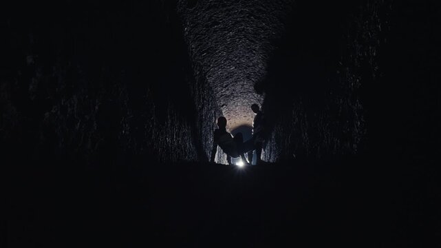 The mysterious silhouettes of several people are seen at the end of a dark passage, backlit by a single light source. They appear to be carrying or attending to a fallen figure in a solemn ceremony.