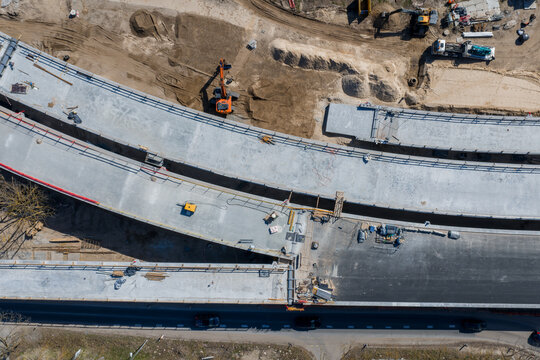 Aerial view shows Southern Bridge phase four in Riga, Latvia, with two curving viaduct spans over a multilane road, crews and machines active near the Daugava corridor.