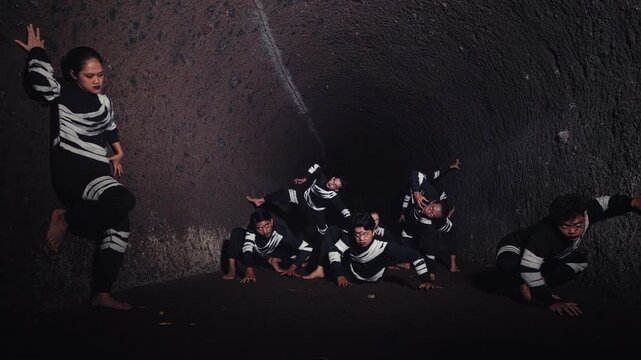 A slow dolly shot reveals a group of performers in unique costumes, frozen in dynamic poses inside a dark cave. The scene depicts a mysterious tribal ritual or a contemporary dance performance.
