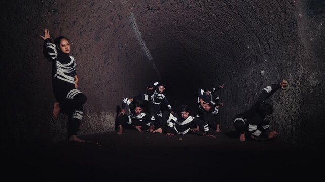 A group of dancers in striking black and white skeleton-like costumes performs a contemporary piece inside a dark tunnel. One woman stands while the men pose low to the ground in a powerful formation.