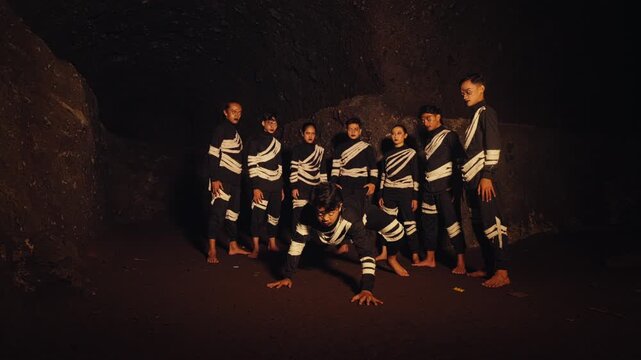 A man in a primal, crawling pose moves toward the camera while his tribe stands behind him with intense expressions. A powerful performance of confrontation or animalistic instinct in a dark cave.