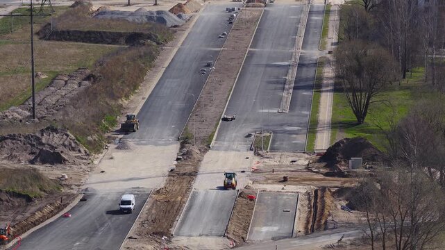 Daytime aerial shows Southern Bridge phase four in Riga, Latvia, with Riga TV Tower, multi lane road, median, path, parkland, and active machinery compacting sand and gravel.