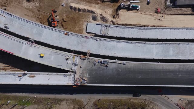 Aerial view of three curved bridge decks under construction in Riga, Latvia, with expansion gaps, temporary railings, an orange excavator, trucks, workers, and paving in bright midday light.