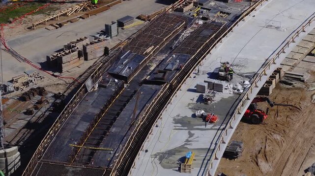 High angle aerial view shows Southern Bridge phase 4 in Riga, Latvia, with curved deck, timber formwork, rebar cages, crane base, telehandler, crews, and wet slab
