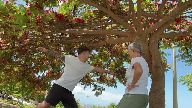A caring young man guides an older woman through a peaceful yoga session outdoors under a beautiful tree. This scene highlights intergenerational connection and the importance of active well-being for