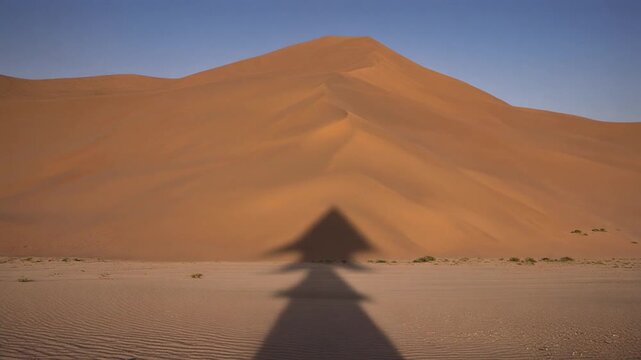 Large triangular shadow of an unseen flying object moving across a rippled desert sand landscape with a huge dune in the background, immersive video footage.
