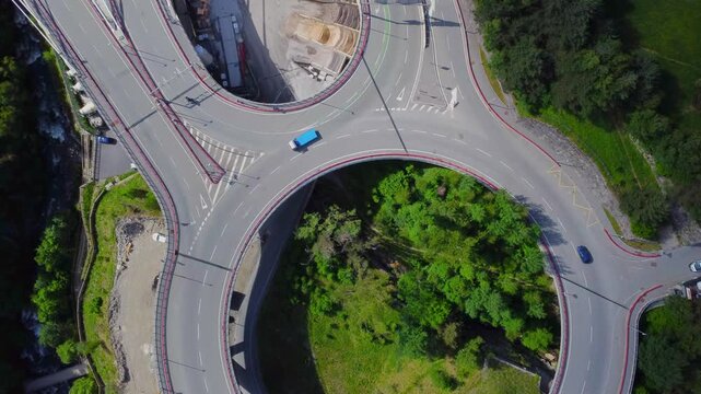 aerial drone shot captures a modern roundabout nestled within the stunning mountainous landscape of Andorra. Vehicles navigate the circular intersection surrounded by lush greenery and the majestic Py