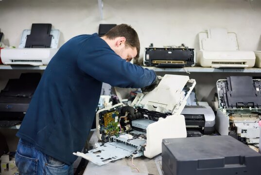 Technician in gloves repairs disassembled printer, surrounded by electronic waste and spare parts.