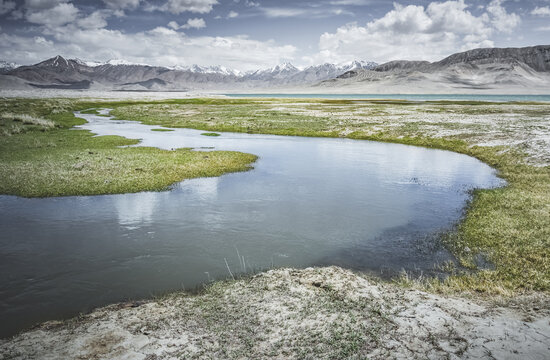 High in the Pamir Mountains of Tajikistan, a river flows along a raft with grassy banks against a backdrop of snow-capped mountain peaks. A panoramic view of the Tien Shan highlands