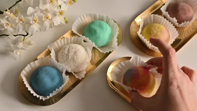 Colorful mochi ice cream balls displayed on elegant gold trays with white paper cups, featuring pastel shades of blue, white coconut, mint green, multicolor swirl, yellow, and purple. A hand gently pi