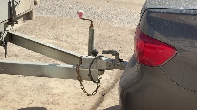 Close-up passenger car hitch trailer in parking lot, sandy ground, metal coupling attached to receiver, safety chain looped