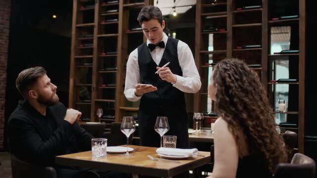 Handheld medium shot of young Caucasian waiter wearing classic outfit taking order on notepad from young couple in elegant formalwear spending romantic date in luxury restaurant