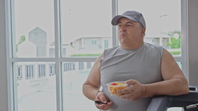 Overweight mature man eating healthy fresh fruit salad after workout near window