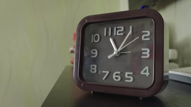 A square, retro-style brown alarm clock sits on a dark shelf, its hands moving to show the passage of time. Concept of waiting, daily schedules, deadlines, and the constant flow of time.