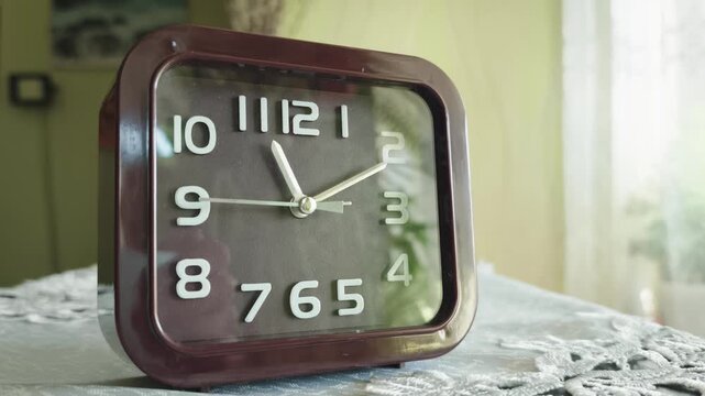 A square analog alarm clock stands on a table with the second hand moving steadily. The dark face with bold white numbers contrasts sharply with the dim interior lighting.