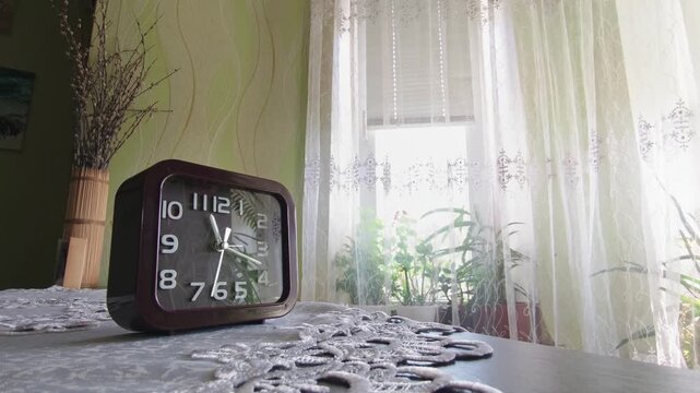 A brown retro alarm clock rests on a table with a lace doily in a bright sunlit room. The daylight streaming through sheer curtains creates a peaceful morning atmosphere.