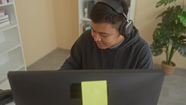 Man wearing headphones writing with pen at computer in a building office, notebook open and monitor visible; focused learning.