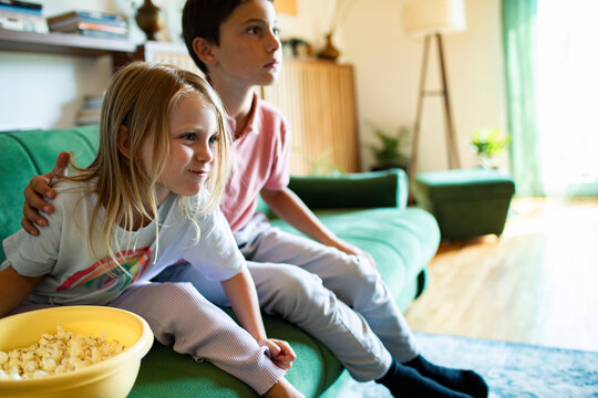 Siblings watching television with popcorn on green couch at home