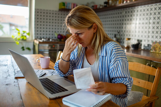 Stressed woman reviewing household bills on laptop at kitchen table
