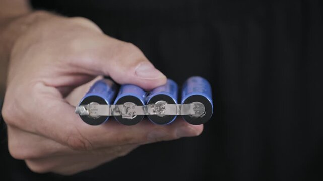 A technicians hand holds and inspects four blue lithium-ion cells connected in series. The spot-welded nickel strip illustrates a DIY power pack assembly.