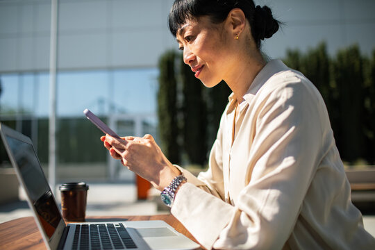 Businesswoman using smartphone and laptop at outdoor cafe