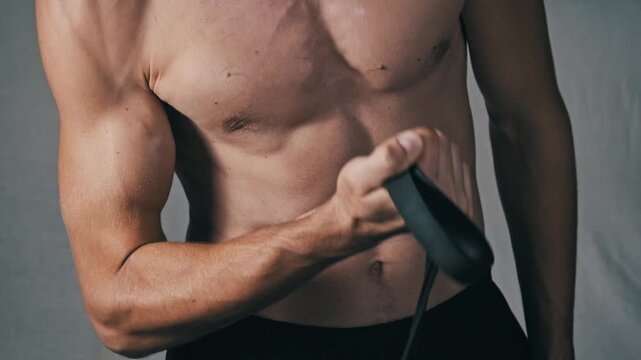 Close-up of a fit mans muscular arm and defined torso performing bicep curls with an elastic band. The shot showcases muscle flexion and tone during strength training.