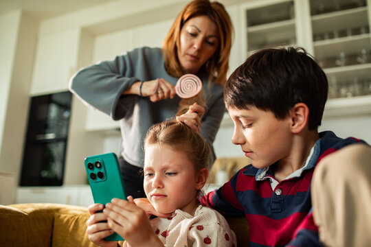 Mother brushing daughter's hair while siblings look at smartphone at home