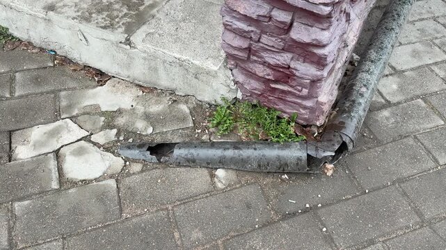Cracked pipe by brick corner leaking onto pavement, corroded metal downspout detached from wall, small hole releasing water, moss