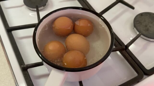 Close-up boiling eggs in iron ladle on gas stove, five brown shells arranged in water, gentle simmer with small bubbles, enamel