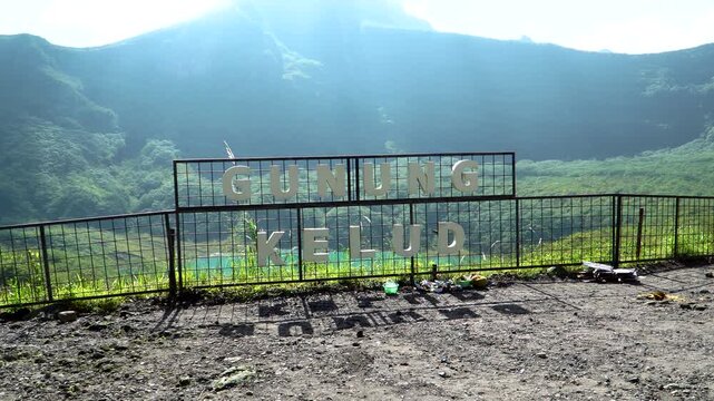 Wide shot of a metal fence displaying a Gunung Kelud sign overlooking the turquoise volcanic crater lake on a bright, sunny day in Kediri, East Java, Indonesia.