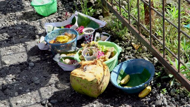 Panning close-up of traditional Javanese ritual offerings with fruits and a coconut placed on the rocky ground by a metal safety fence at Mount Kelud crater, Kediri, East Java, Indonesia.