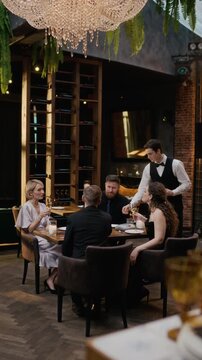 Vertical wide shot of young Caucasian waiter serving dinner to cheerful couple friends raising glasses and celebrating joyous event in luxury restaurant together