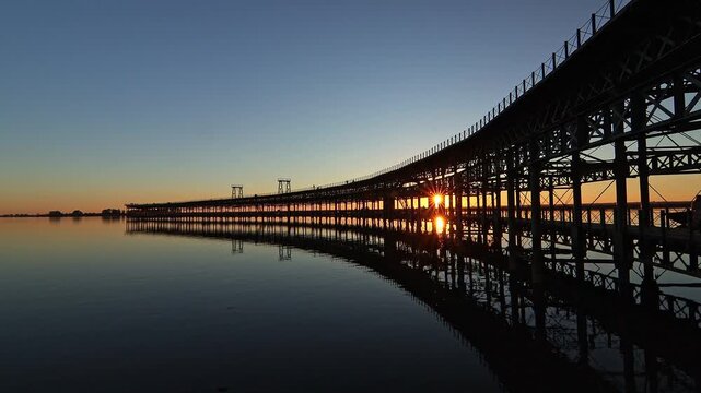 Stunning silhouette of the historic iron pier reflected on calm water. A sharp sunstar glows through the industrial frame at dusk. Ideal for travel, history, and architectural projects.