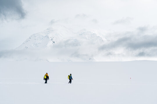 View of two explorers with backpacks walking across a vast snowfield towards a massive snow-covered mountain under a cloudy sky Antarctica.