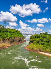 Obraz premium Breathtaking panoramic view of the majestic Iguazu Falls with the river flowing through a lush green canyon and a tourist boat under a clear blue sky with fluffy white clouds