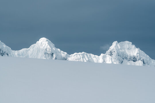 View of snow-covered mountain peaks rising above a smooth white slope under a dark overcast sky Antarctica.