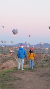 A couple stands hand in hand, gazing at hot air balloons rising in the tranquil dawn sky of Cappadocia, Turkey. The stunning landscape offers a magical setting for unforgettable moments.