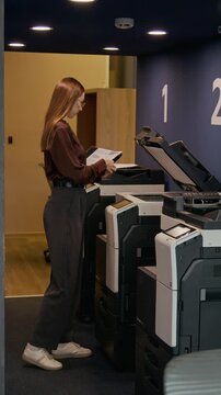 Vertical shot of young woman walking in office and using multifunctional printer to copy or scan business document