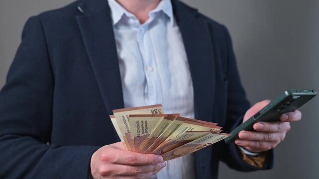 A middle-aged male with light skin is holding a large stack euro of cash in one hand and checking his smartphone with the other hand