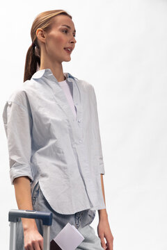 Young woman stands holding a suitcase handle and travel documents, wearing a casual white shirt and light pants. Concept of travel, vacation, and preparation for journey.