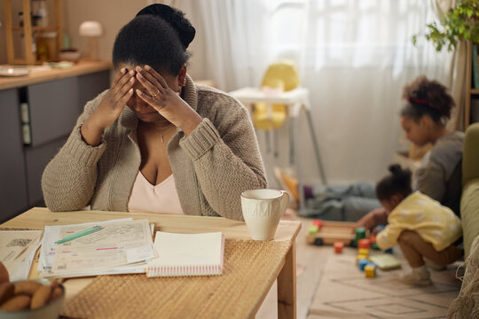 African American mother covering face while reviewing household bills, struggling with financial stress as children playing nearby. Ideal for poverty, debt, budgeting, family hardship themes