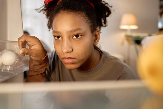 Portrait of Black teenage girl looking into nearly empty refrigerator, holding egg tray and expressing food insecurity. Useful for poverty, hunger, cost of living, social aid campaigns