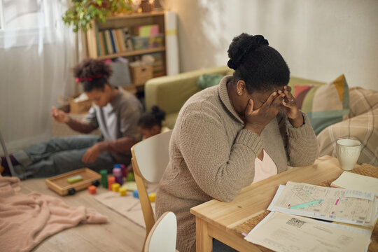 African American mother covering face while sitting by unpaid bills, children playing behind her at home. Useful for poverty, financial stress, debt, family hardship marketing