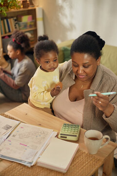 Vertical shot of Black mother holding toddler while calculating household bills, with older child sitting nearby, showing financial hardship. Ideal for poverty, budgeting, social support campaigns