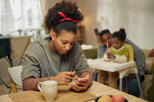 Black teenage girl checking phone while eating at table, mother caring for child in background, showing financial stress and low income parenting. Useful for poverty, family budget, welfare support