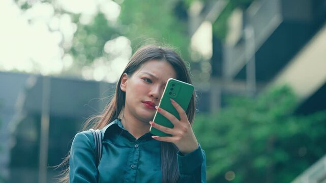 Frustrated Asian woman in a teal satin shirt stands outdoors, frowning while looking at her smartphone. She is feeling stressed by fake social media content, AI misinformation, and online scams.