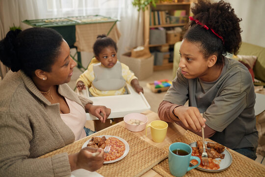African American mother and teenage daughter eating modest meal with toddler nearby, discussing family hardship at home. Useful for poverty, food insecurity, low income household campaigns