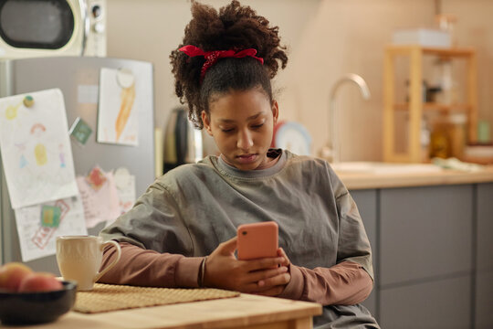 Teenage girl using smartphone while sitting at kitchen table, checking messages with worried expression and reflecting financial hardship. Useful for poverty, family stress, social support themes