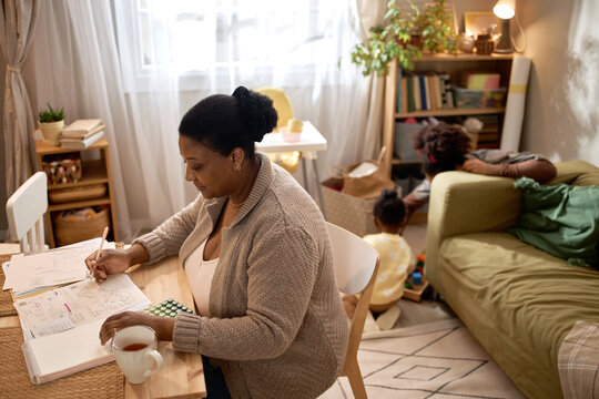 African American mother managing household bills while children playing in background at home. Ideal for poverty, financial stress, single parenting, budgeting, social support campaigns